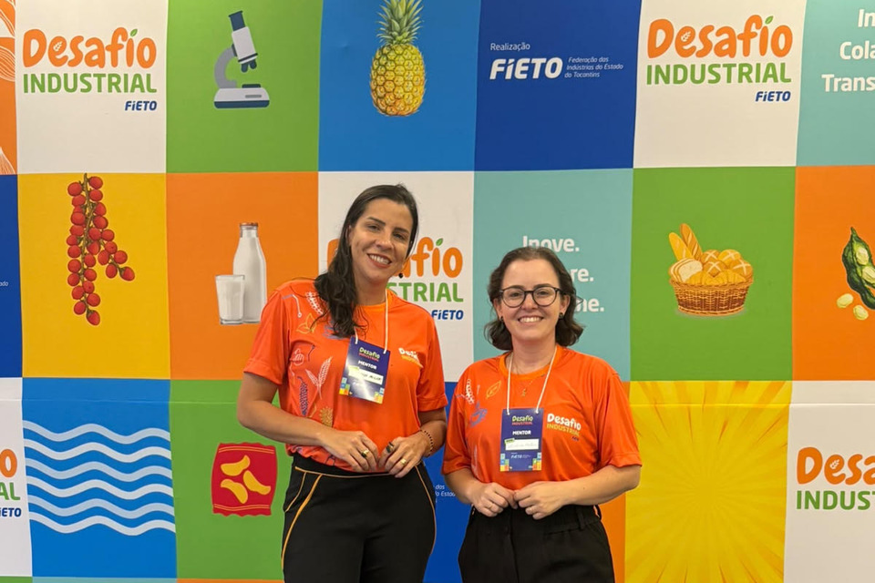 Fotografia colorida de duas mulheres em pé, lado a lado, usando camisetas laranjas do Desafio Industrial FIETO e crachás de mentoras. Ao fundo, há um painel colorido com elementos relacionados à indústria de alimentos e a identificação do evento.